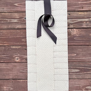 May include: A white rectangular rug with a black ribbon tied in a bow at the top. The rug has a textured surface and is made of a woven material.
