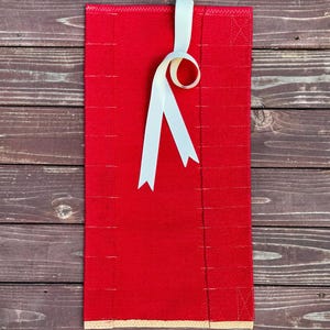 May include: A rectangular red fabric item with a white ribbon tied at the top. The fabric has a textured appearance and a beige border at the bottom. The ribbon is looped and has two pointed ends. The background is a dark brown wooden surface.