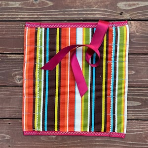 May include: A colorful striped placemat with a pink ribbon tied in a bow on top. The placemat has a scalloped edge and is made of a woven material.
