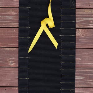 May include: A rectangular black placemat with a ribbed texture and gold stitching. A bright yellow ribbon is tied in a knot at the top. The placemat is displayed on a wooden surface.