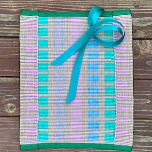 May include: A woven placemat with a checkered pattern in shades of pink, blue, and green. The placemat has a green border and a teal ribbon tied in a bow on top.