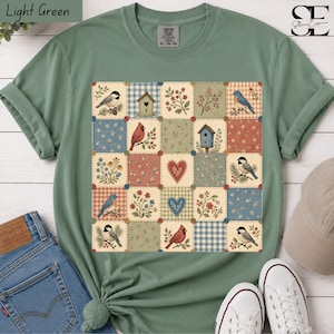 Può includere: Una t-shirt verde chiaro con una stampa grafica centrale in stile patchwork. Il design è composto da pannelli quadrati che mostrano illustrazioni di uccelli, casette per uccelli, cuori e motivi floreali nei toni del blu, rosso e crema.