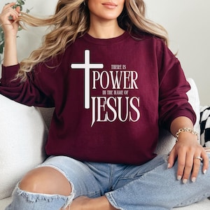 Puede incluir: Una sudadera color burdeos con una cruz blanca y el texto "There is power in the name of Jesus".