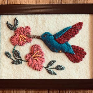 May include: Framed textile art featuring a hummingbird with blue body and red wings, hovering near pink flowers with yellow stamens. The artwork is set against a cream-colored background and framed in a dark brown wooden frame.