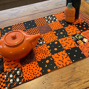 May include: A rectangular patchwork table runner featuring a variety of orange and black fabric squares with patterns including bats, pumpkins, polka dots, and owls. A solid orange ceramic teapot sits on the left side of the runner.