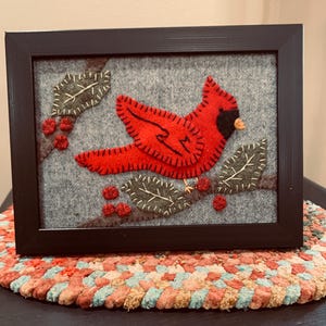 May include: Framed felt art featuring a vibrant red cardinal perched on a branch with green leaves and red berries. The bird is set against a gray background and framed in a black rectangular frame. The artwork is displayed on a colorful braided mat.