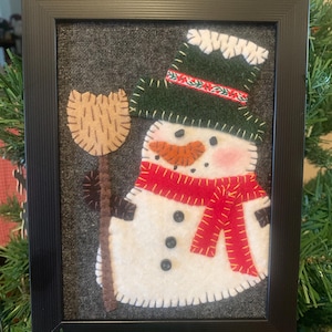 May include: A framed felt applique of a white snowman wearing a dark green top hat with a red and white floral band, a bright red scarf with yellow stitching, and three black buttons. The snowman holds a tan broom against a dark gray background.