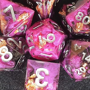 May include: A set of polyhedral dice made from translucent resin with a swirling magenta and black pigment, embedded with metallic copper flakes. Each die features sharp edges and large, deeply engraved numbers painted in a bright silver finish.