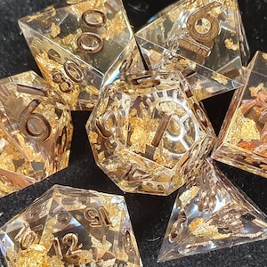 May include: A set of seven clear resin dice with gold leaf inclusions and brown numbering. The dice are in various polyhedral shapes, including a d4, d6, d8, d10, d12, d20, and a percentile die. Ideal for tabletop gaming.