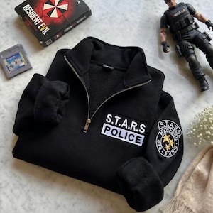 Könnte beinhalten: Schwarzes Sweatshirt mit Reißverschluss, "S.T.A.R.S. POLICE" Aufnäher und rundem Sternenaufnäher. Das Sweatshirt hat einen hohen Kragen und einen silbernen Reißverschluss. Eine Resident Evil-Spielbox und eine Figur sind ebenfalls im Bild.