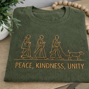 Può includere: T-shirt verde oliva con scollo tondo. La maglietta presenta un disegno ricamato di tre figure che camminano con un cane, con le parole "PEACE, KINDNESS, UNITY" sotto. Il disegno è di colore giallo dorato.