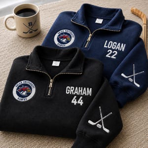Op de afbeelding: Twee marineblauwe en zwarte sweatshirts met een kwartrits, geborduurde namen en nummers en een Briar University Hawks-logo. Er is ook een beige mok met een hockeysticklogo aanwezig.