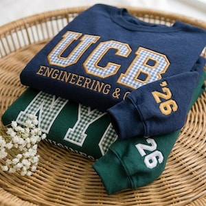 Pode incluir: Camisolas azul marinho e verde com texto bordado. A camisola azul marinho apresenta "UCB ENGINEERING & C" com um padrão xadrez. A camisola verde tem "NY" com um padrão xadrez. As mangas exibem o número "26".