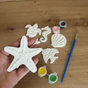 May include: A collection of white, sea-themed craft items, including a starfish, seahorse, turtle, fish, and seashell. Small paint pots with yellow, green, pink, and black paint are included, along with a blue paintbrush. The items are arranged on a wooden surface.