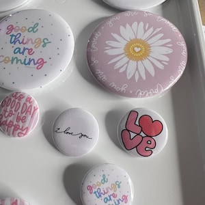 Può includere: Una collezione di bottoni rotondi con vari disegni e testo. Alcuni bottoni presentano frasi come "Good things are coming" e "I love you". Altri mostrano una margherita, la parola "Magic" e la parola "Love" con un cuore.