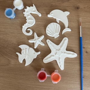 Puede incluir: Un kit de manualidades con formas blancas de criaturas marinas: caballito de mar, delfín, estrella de mar, pez y concha. El kit incluye botes de pintura en azul, blanco, rojo y naranja, además de un pincel. Los artículos están dispuestos sobre una superficie de madera.