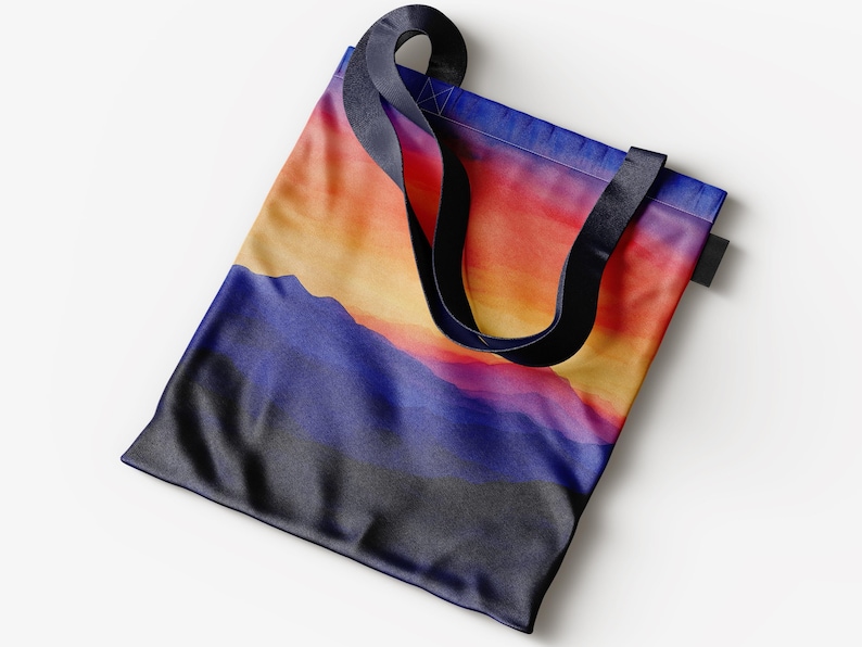 K&ouml;nnte beinhalten: Eine Tragetasche mit schwarzen Henkeln und einem farbenfrohen Sonnenuntergangs-Landschaftsdruck. Die Tasche zeigt einen Farbverlauf von Orange, Gelb, Rosa und Lila mit einem dunkelblauen Gebirgszug im Vordergrund.