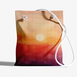 K&ouml;nnte beinhalten: Eine wei&szlig;e Canvas-Tragetasche mit einem auf die Vorderseite gemalten Sonnenuntergang. Der Sonnenuntergang ist ein Farbverlauf von Orange, Rot und Lila mit einem wei&szlig;en Kreis in der Mitte.