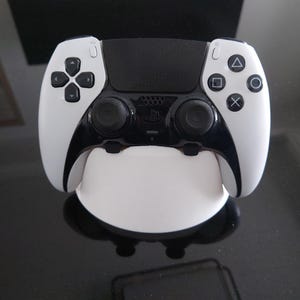 Könnte beinhalten: Ein weiß-schwarzer PlayStation 5 DualSense Wireless-Controller. Er hat einen schwarzen Mittelteil mit zwei Analogsticks und eine weiße Außenschale. Mit Aktionstasten und einem Steuerkreuz.