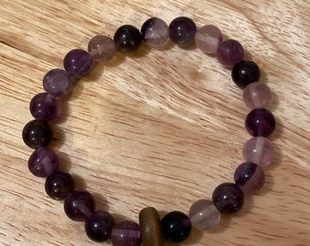 May include: A purple and white gemstone bracelet with a wooden bead accent. The bracelet is made of round beads and is strung on a clear elastic cord.