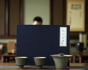 May include: A tea set with two small cups and a lidded gaiwan, all in a muted green color with gold rims. The set is arranged on a wooden surface in front of a dark blue box with text on a white label. The background is a room with wooden furniture.