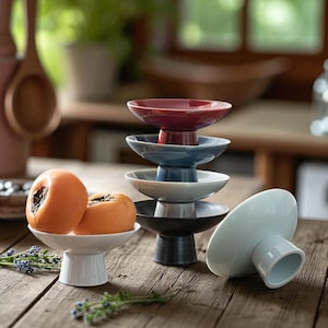 May include: A selection of small, pedestal-style ceramic bowls in shades of red, blue, white, and black. Two orange persimmons sit on a white bowl. The bowls are displayed on a rustic wooden surface.