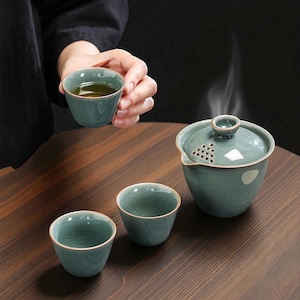 May include: A teal ceramic tea set comprising a teapot, two teacups, and one teacup being held. The teapot features a lid with a small handle and a spout with small holes. The teacups have a gold rim. The tea set is on a wooden table.