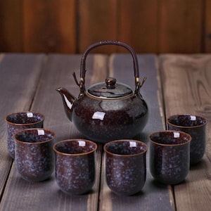 May include: A dark brown ceramic teapot with a woven handle and a matching lid, accompanied by six small, speckled cups. The tea set is arranged on a wooden surface, showcasing a traditional tea ceremony aesthetic.