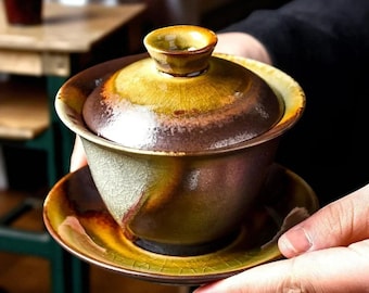 May include: A ceramic tea set with a lid, cup, and saucer. The set features a crackle glaze in shades of brown, yellow, and green. The tea cup is held in someone's hands. The tea set is designed for tea ceremonies.