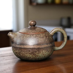 May include: A ceramic teapot with a rounded body, a curved handle, and a small spout. The teapot has a speckled, mottled appearance with shades of brown, green, and bronze. A small, round knob sits atop the lid.