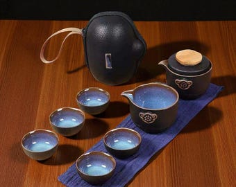 May include: A ceramic tea set with a black carrying case. The set includes a teapot, a pitcher, and six small cups. The cups and pitcher have a blue and brown glaze. The teapot has a wooden lid. The set is on a blue cloth.