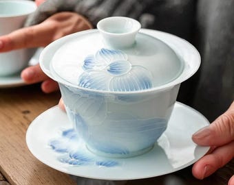May include: A white porcelain gaiwan tea set with a matching lid and saucer. The set features a delicate blue floral design. The gaiwan is designed for brewing and serving tea, and is held by a person.