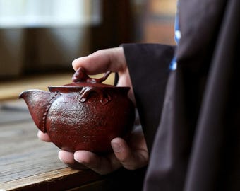 May include: A small, reddish-brown ceramic teapot with a detailed design. The teapot has a curved spout, a decorative lid, and a textured surface. The teapot is held in a person's hand, suggesting its size and craftsmanship. A traditional tea accessory.