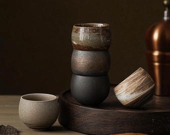 May include: A stack of three handcrafted ceramic cups in earthy tones of brown, gray, and black. One cup is tilted on a wooden tray, and another cup sits separately. The cups have a rustic, textured finish.
