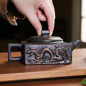 May include: A dark brown, square teapot featuring a dragon relief. The teapot has a small handle and spout, and a lid with a small square handle. It sits on a wooden surface. The teapot is likely made of ceramic.