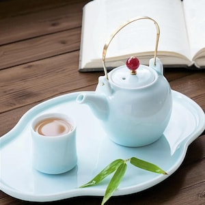 May include: A light blue ceramic tea set with a teapot, cup, and serving tray. The teapot has a gold handle and a red knob on the lid. The cup is filled with tea. The set is on a wooden surface.