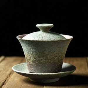 May include: A ceramic gaiwan tea set, comprising a lidded cup and saucer. The set features a textured, speckled finish in green, brown, and pink hues. The cup has a flared shape, and the saucer is low-profile. The background is dark.