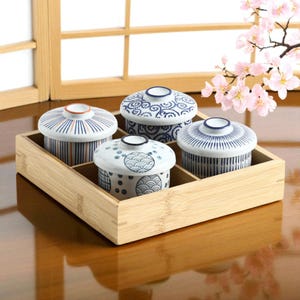 May include: Four small, lidded ceramic bowls with blue and white patterns sit in a bamboo tray. The bowls have various designs, including stripes and swirls. The tray is rectangular with divided sections. Cherry blossoms are in the background.