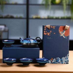May include: A ceramic tea set with a blue and black gradient glaze. The set includes a teapot and four matching teacups. The set is displayed with a decorative box featuring a crane and floral design. The tea set is presented on a dark blue surface.