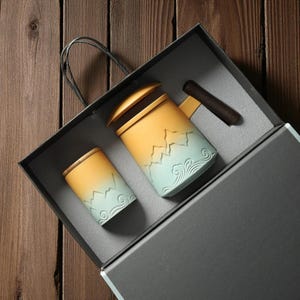 May include: A ceramic tea set, comprising a mug and a cup, presented in a gift box. The set features a yellow and blue gradient design with mountain and wave motifs. The mug has a wooden handle and lid.