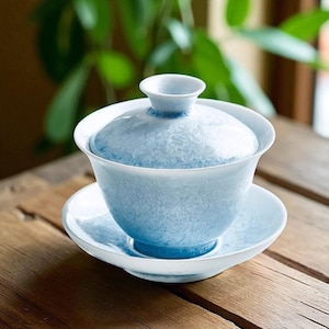 May include: A light blue ceramic gaiwan tea set with a matching lid and saucer. The gaiwan has a textured surface and a white rim. The set is displayed on a wooden table, with green foliage in the background.