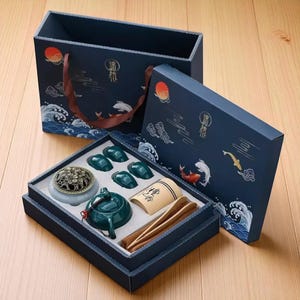 May include: A dark blue gift set with a decorative design featuring waves, fish, and a red sun. The set includes a teapot, cups, incense burner, and other accessories. The box has a matching design and a brown handle.