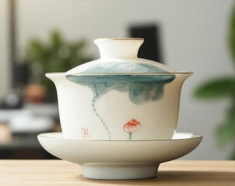 May include: A white porcelain tea set comprising a lidded cup and saucer. The cup and lid are decorated with a blue and green lotus design and gold trim. A red flower is painted on the cup. The set sits on a wooden surface.