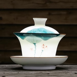 May include: A white ceramic gaiwan tea set with a matching lid and saucer. The gaiwan features a hand-painted design of a blue lotus leaf and pink flowers. The rim of the gaiwan and saucer have a brown trim.