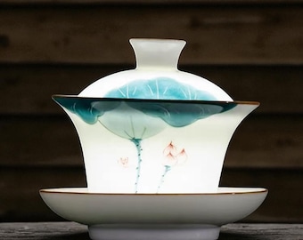 May include: A white ceramic gaiwan tea set with a matching lid and saucer. The gaiwan features a hand-painted design of a blue lotus leaf and pink flowers. The rim of the gaiwan and saucer have a brown trim.