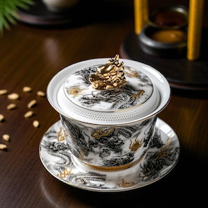 Peut inclure: Service à thé en céramique blanche avec un motif de dragon noir et or. L'ensemble comprend une tasse, une soucoupe et un bol couvert avec une figurine de dragon doré. Posé sur une surface en bois sombre.