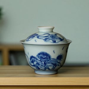 Peut inclure: Bol à thé en céramique blanche avec couvercle, orné d'illustrations bleues de créatures mythiques et de motifs tourbillonnants. Le bol a une forme arrondie et une petite poignée sur le couvercle. Le design rappelle l'art asiatique traditionnel.