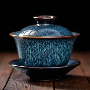 May include: A blue ceramic gaiwan tea set with a matching lid and saucer. The gaiwan has a textured surface with a dark brown rim. The set is designed for traditional tea ceremonies.