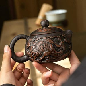 May include: A dark brown ceramic teapot with detailed dragon relief carvings. The teapot has a curved handle, a spout, and a domed lid with a small knob. The dragon design adds a decorative element to the tea set.