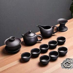 May include: A black ceramic tea set, comprising a teapot, cups, a creamer, and a strainer. The set is displayed on a light wood surface, against a dark grey backdrop. The tea set is designed for serving tea.